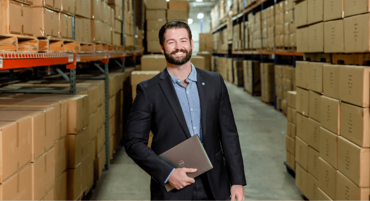 A man in a suit stands smiling in a warehouse aisle lined with shelves stacked with cardboard boxes, holding a closed laptop in his right hand—ready to streamline warehousing and fulfillment operations.
