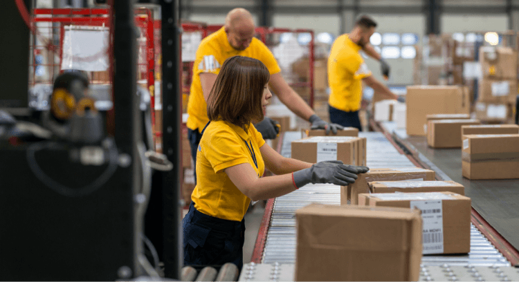 Warehouse workers in yellow shirts efficiently sort and pack cardboard boxes on a conveyor belt, showcasing streamlined fulfillment processes amid shelves and stacks in the busy warehousing area.