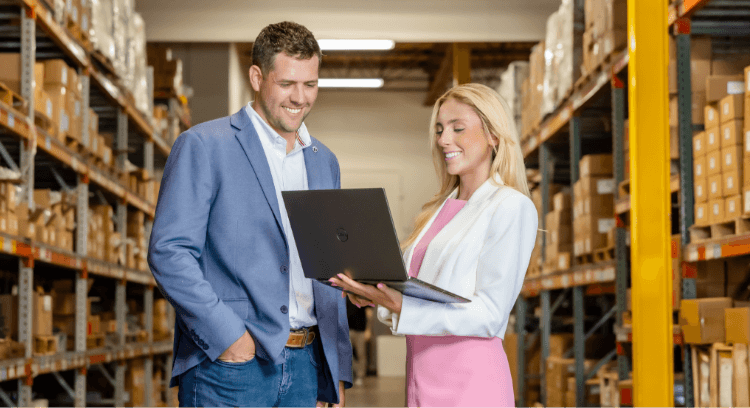 Two business professionals, a man in a blue suit and a woman in a white blazer, stand smiling and looking at a laptop in a fulfillment warehouse filled with shelves and boxes.