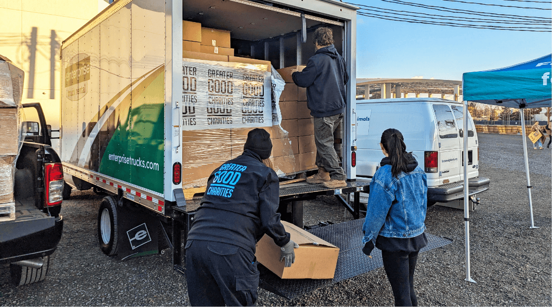 Three people unload cardboard boxes from the back of a delivery truck, stacking them on the ground near other vehicles in a parking lot on a sunny day, as part of their warehousing and fulfillment operations.