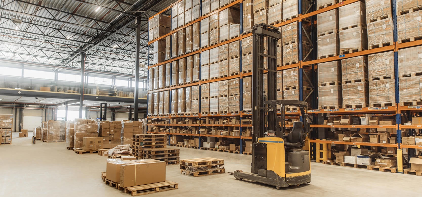 A well-lit warehouse interior designed for warehousing and fulfillment, featuring tall shelves stacked with boxed goods, a yellow forklift, and several pallets and boxes on the floor beneath high ceilings and large windows.