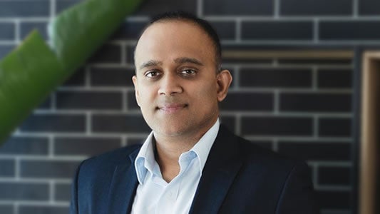 A man in a dark suit and white shirt stands before a dark brick wall, looking at the camera with a neutral expression. On the left, a green plant adds contrast—perfect for announcing FreightWaves' 2026 FreightTech 100 List achievements.