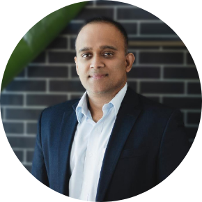A man in a dark suit jacket and white shirt stands in front of a brick wall, looking at the camera with a slight smile.