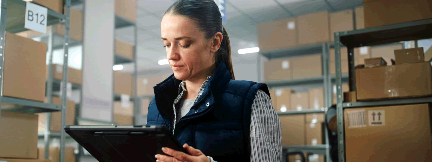 A woman in a warehouse stands among shelves of cardboard boxes, looking at a tablet. She wears a striped shirt and dark vest, focused on managing Large Parcel Shipping in this organized storage area.