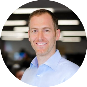 A man with short light brown hair, wearing a light blue collared shirt, smiles at the camera. The background is blurred, showing office lights and indistinct people.