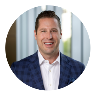A man in a blue checkered suit jacket and white shirt smiles in front of a blurred background with vertical lines.