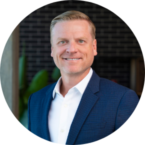 A smiling man wearing a blue blazer and white shirt stands in front of a dark brick wall with green plants in the background.