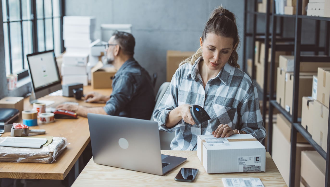 A woman scans a package with a barcode scanner at a desk with a laptop, using shipping tools, while a man works at another desk in a room filled with cardboard boxes and shelves.