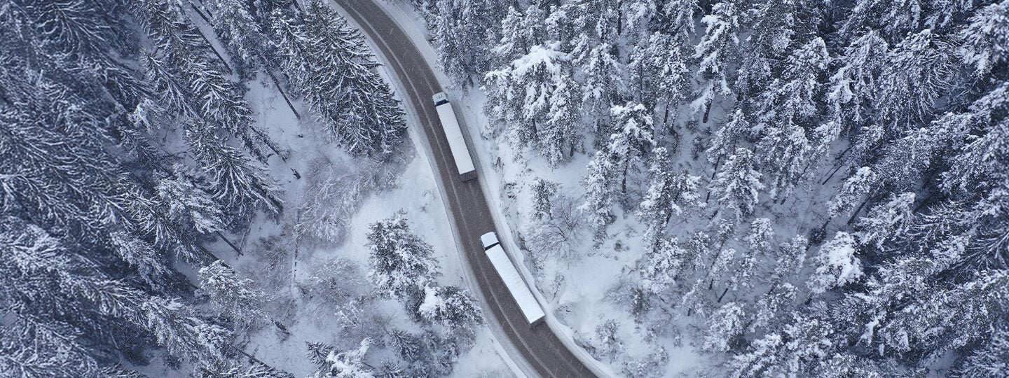 Aerial view of two trucks, possibly transporting UPS Oversized Package shipments, driving on a winding road through a dense, snow-covered forest in winter.