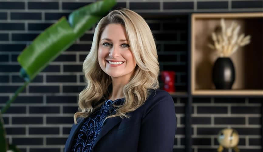 A smiling woman with long blonde hair wearing a dark blazer stands indoors. Behind her, a black brick wall with shelves holding decorative items, including a vase with dried plants, sets the scene—perfect for a third-party logistics office space. A green leaf is in the foreground.