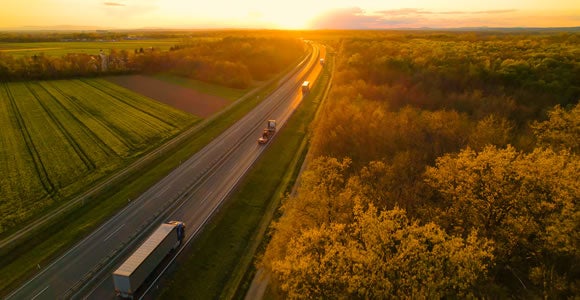 Aerial view of a highway with trucks, including refrigerated freight vehicles, and cars driving through green fields and trees at sunset, with golden sunlight illuminating the landscape.