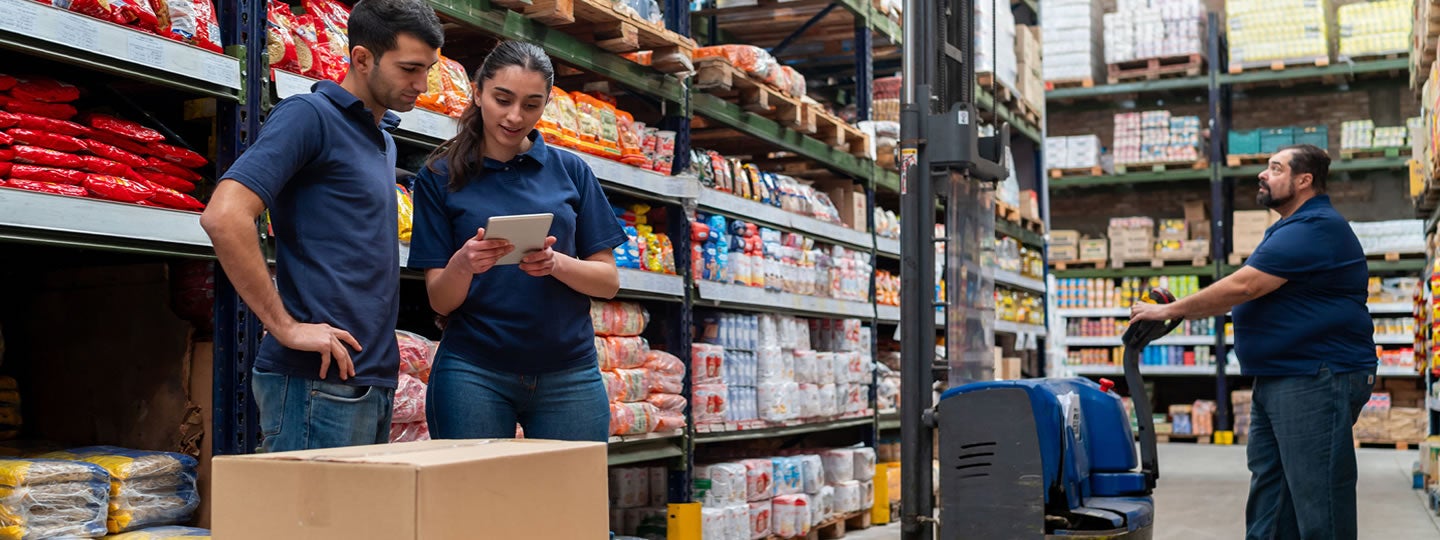 Three warehouse workers in blue shirts organize inventory; two review information on a tablet, while another operates a forklift in an aisle lined with shelves of packaged goods and boxes.
