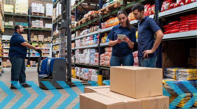 Three workers in a warehouse stand near stacked boxes; two review information on a tablet while another operates equipment among shelves stocked with various goods.