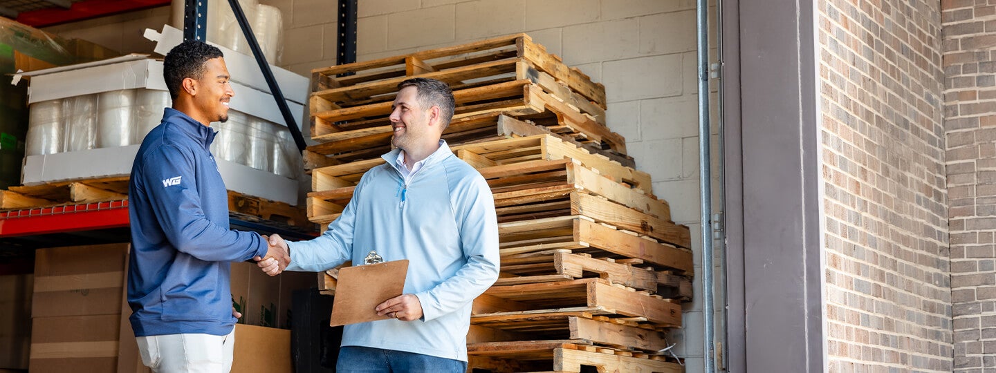 Two men are standing in a warehouse, shaking hands and smiling. One is holding a clipboard, and stacks of wooden pallets are visible in the background. Light is coming in from an open doorway on the right.