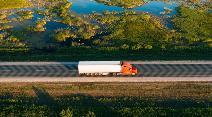 An aerial view of a red semi-truck with a white trailer driving on a rural road, surrounded by green grass and wetlands with water patches.