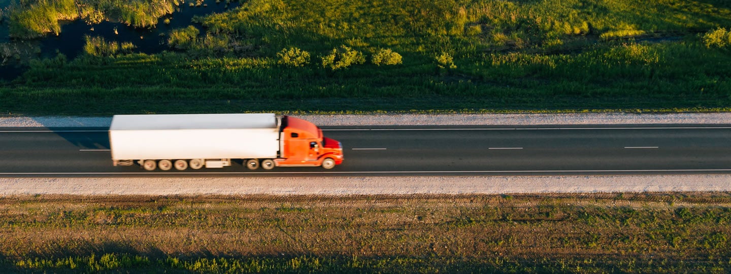 Aerial view of an orange semi-truck with a white trailer driving on a straight, empty highway bordered by green grass and a small body of water.