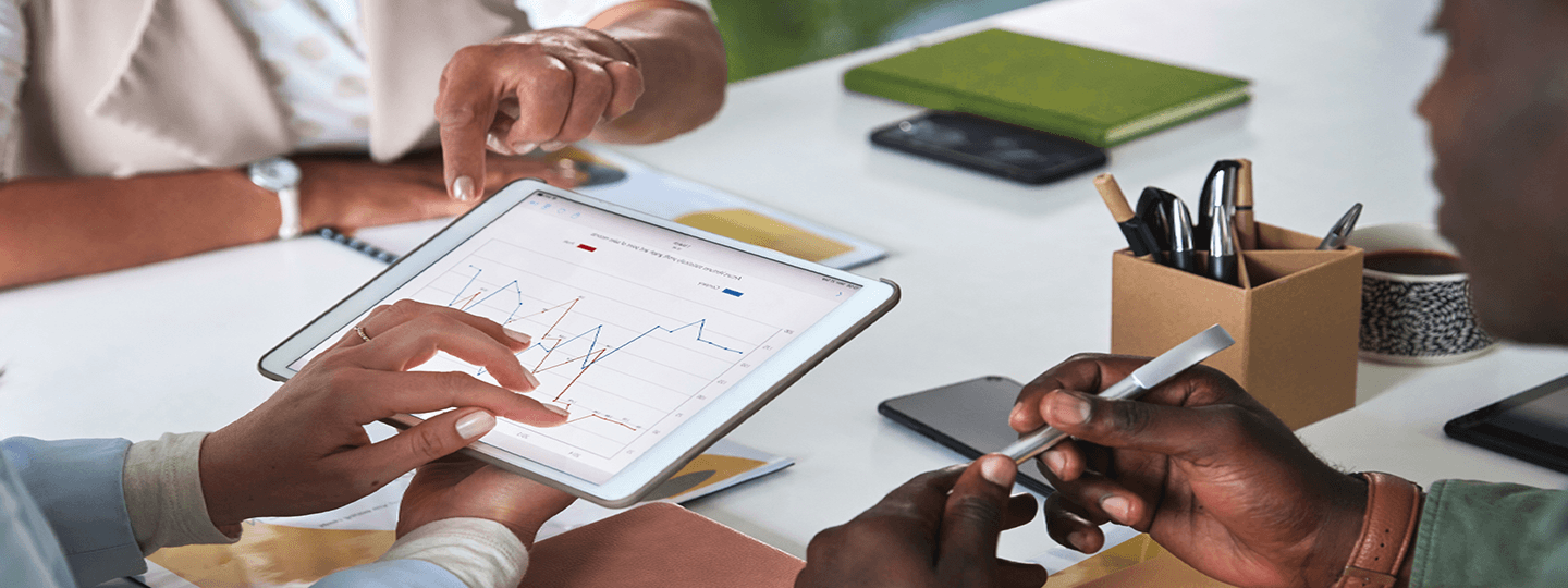 Two people sit at a table discussing a tablet displaying a line graph. One person points at the screen while the other holds a stylus, with notebooks, pens, and a smartphone nearby.