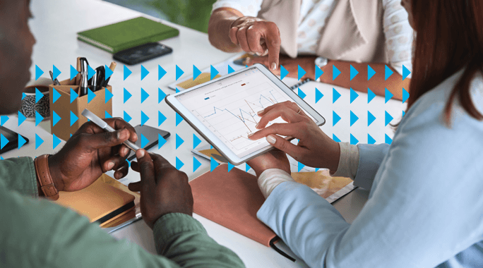 Three people sit at a table discussing data on a tablet displaying a line graph. Notebooks, phones, and papers are on the table, and one person gestures towards the screen while another holds a pen. Blue arrow pattern overlays the image.