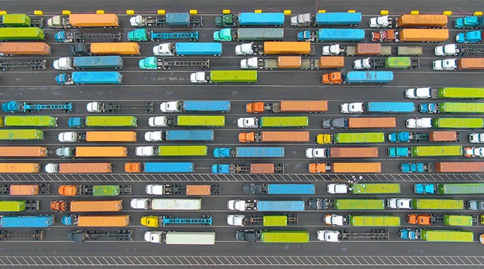 Aerial view of rows of colorful trucks and cars, some likely carrying a Hazmat Bill of Lading, lined up in multiple lanes on a road, creating a vibrant pattern of orange, blue, green, and white vehicles.