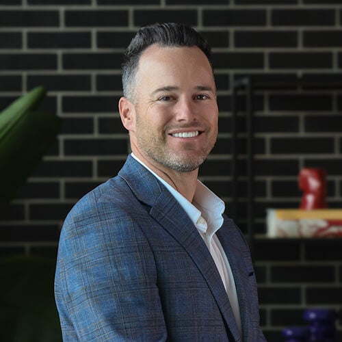 A man with short dark hair and a trimmed beard, wearing a blue plaid suit jacket and white shirt, smiles while standing in front of a black brick wall with shelves and decorative items in the background.