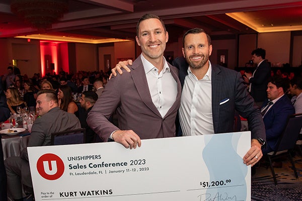 Two smiling men in suits stand together at a crowded event, holding a large check for $1,200 made out to Kurt Watkins at the Unishippers Sales Conference 2023 in Fort Lauderdale, Florida.