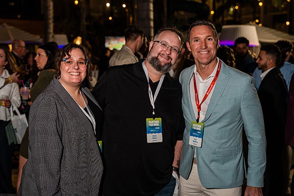 Three people wearing event badges smile and pose together at an outdoor evening event with a crowd of people and string lights in the background.
