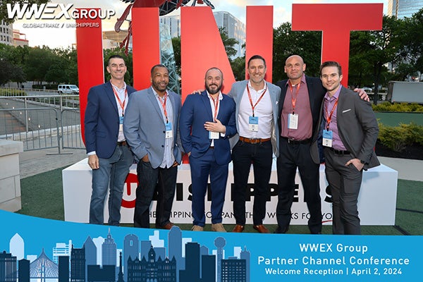 Six men in business attire stand smiling in front of a large red UNT sculpture outdoors. They wear conference badges and lanyards. A banner at the bottom reads WWEX Group Partner Channel Conference, April 2, 2024.