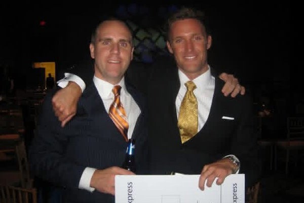 Two men in suits, one holding a drink and the other holding a rectangular white sign, stand side by side with arms around each other, smiling at the camera in a dimly lit setting.