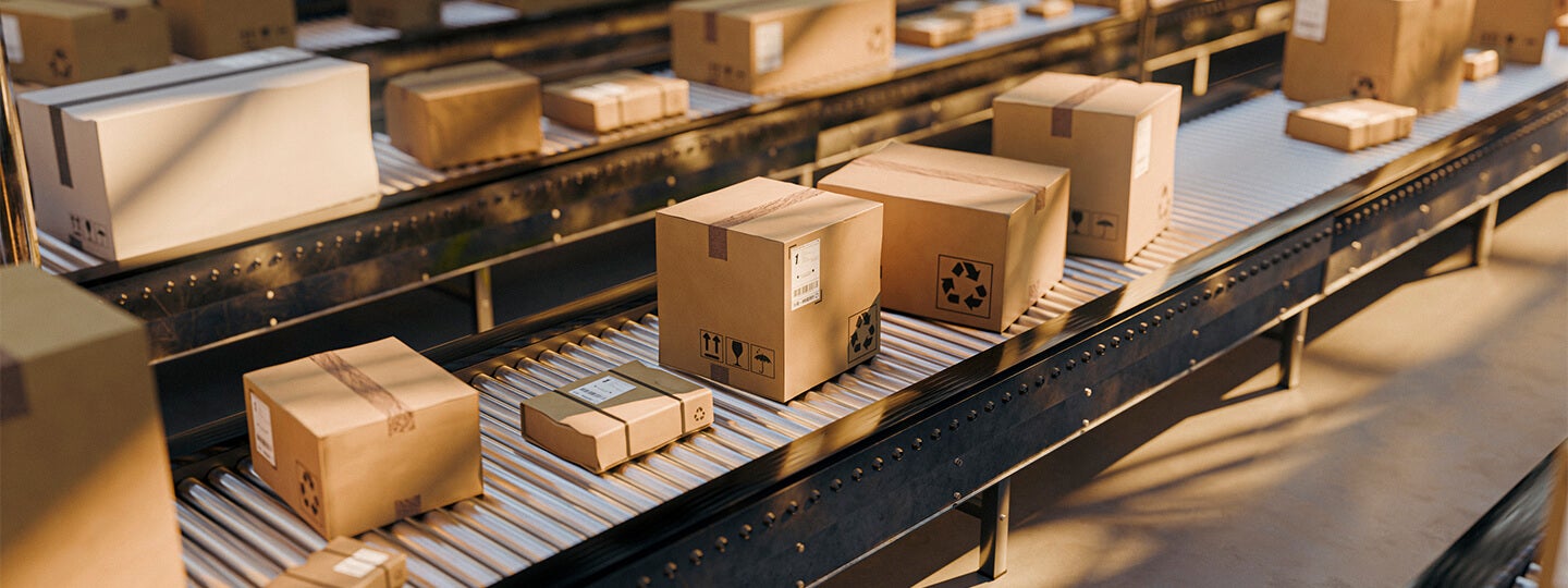 Cardboard boxes of various sizes move along a conveyor belt in a warehouse, illuminated by warm lighting. The scene highlights packaging and shipping as part of an efficient manufacturing supply chain operation.