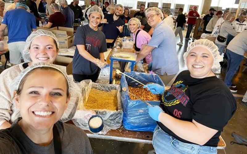 A group of smiling volunteers wearing hairnets and gloves work together to package food at a busy indoor event. Tables are filled with containers of food ingredients and packaging materials.