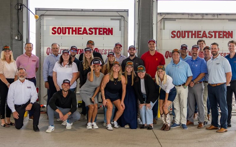A group of people pose and smile in front of two large trucks labeled SOUTHEASTERN. Most are wearing casual or business-casual clothing and matching hats, and the setting appears to be a warehouse or loading dock.