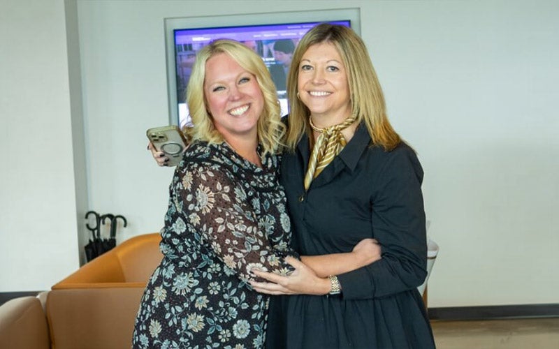 Two women smiling and hugging indoors. One wears a floral dress and holds a phone, the other wears a dark coat with a gold scarf. They stand in front of a brown sofa and a TV screen displaying a website in the background.
