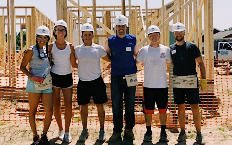 Six people wearing hard hats and construction gear stand in front of a wooden house frame at a construction site, smiling for the camera. Orange safety fencing and building materials are visible in the background.