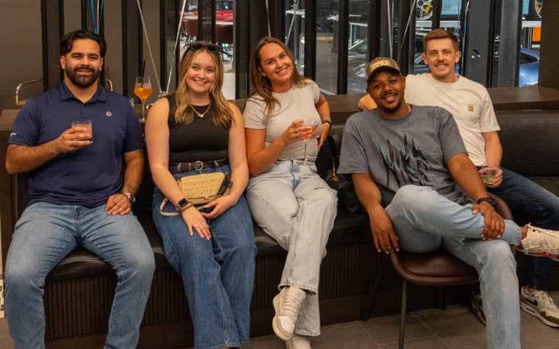 Five people sit on a bench indoors, smiling at the camera and holding drinks. They are casually dressed and appear relaxed, enjoying a social gathering.