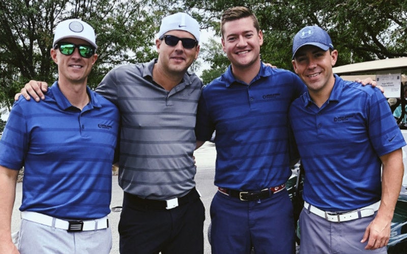 Four men stand together outdoors, smiling for the camera. Three wear matching blue golf shirts and white belts, while one wears a gray striped shirt and a cap. Trees and a golf cart are visible in the background.