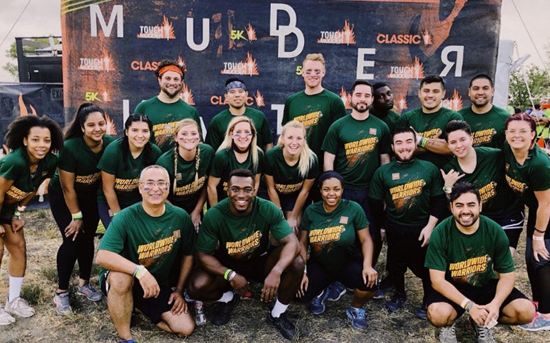 A group of people in matching green “Moulding Warriors” shirts pose and smile together outdoors in front of a Tough Mudder obstacle course sign. Some crouch in front while others stand behind them.