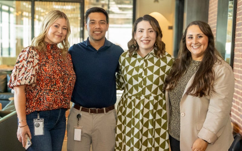 Four people stand side by side indoors, smiling at the camera. Three women and one man are dressed in business casual attire, with office lanyards visible on two of them. The background shows a modern office setting.