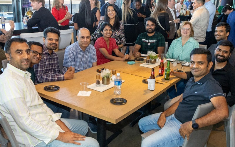 A group of ten people sit and smile around a table in a lively indoor setting, with drinks and small plants on the table. Other groups are visible socializing in the background.