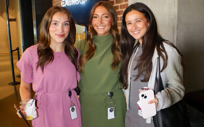 Three women stand together smiling indoors. They wear business-casual outfits and ID badges around their waists. One holds a drink, another holds a canned beverage and phone. A brick wall and signage are in the background.