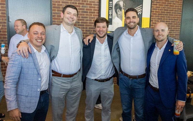 Five men wearing suits and white shirts smile together indoors, standing close with their arms around each other, in front of a brick wall. Two of them hold drinks, and another man is visible in the background.