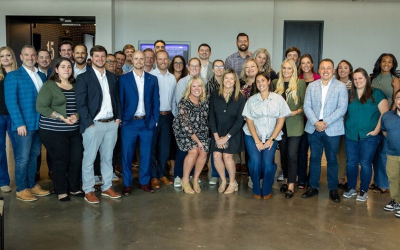 A large group of professionally dressed adults stands closely together indoors, smiling at the camera against a neutral background with modern decor and a wall-mounted screen.