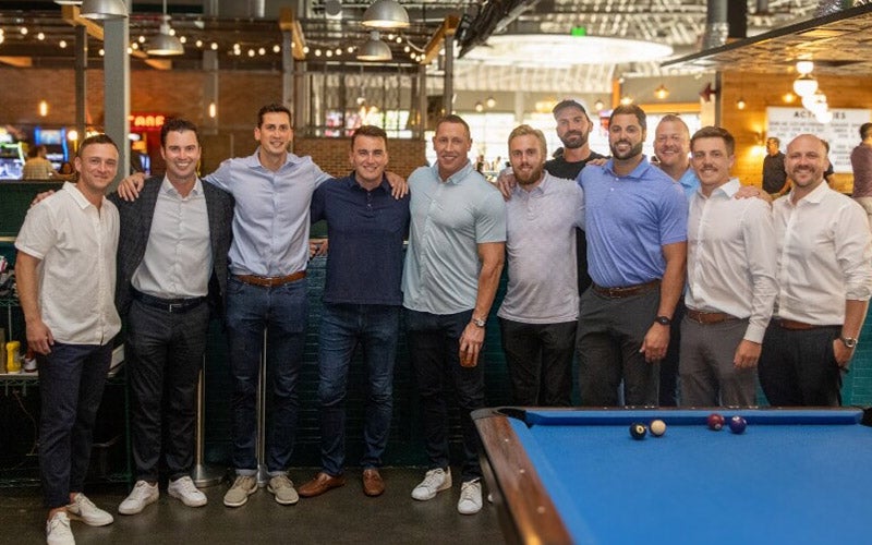 A group of eleven men stand together and smile for a photo in a casual indoor setting near a blue pool table. Most are wearing collared shirts and slacks. The background features bright lights and a lively atmosphere.