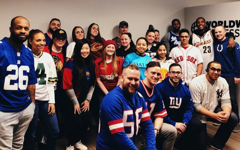 A group of people pose together indoors, smiling and wearing various sports jerseys, including football, baseball, and hockey teams. A sign reading WORLDWIDE is partially visible on the wall behind them.
