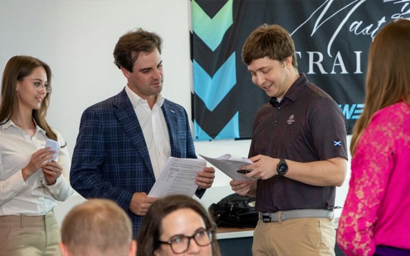 Four people stand and converse while looking at documents during a professional event. Two men are in business casual attire, and two women are present, one in a pink blouse and one in a white shirt. A banner hangs in the background.
