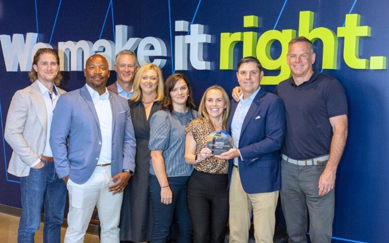 A group of eight professionally dressed people stand smiling in front of a blue wall with the words we make it right. Two people in the center are holding an award.