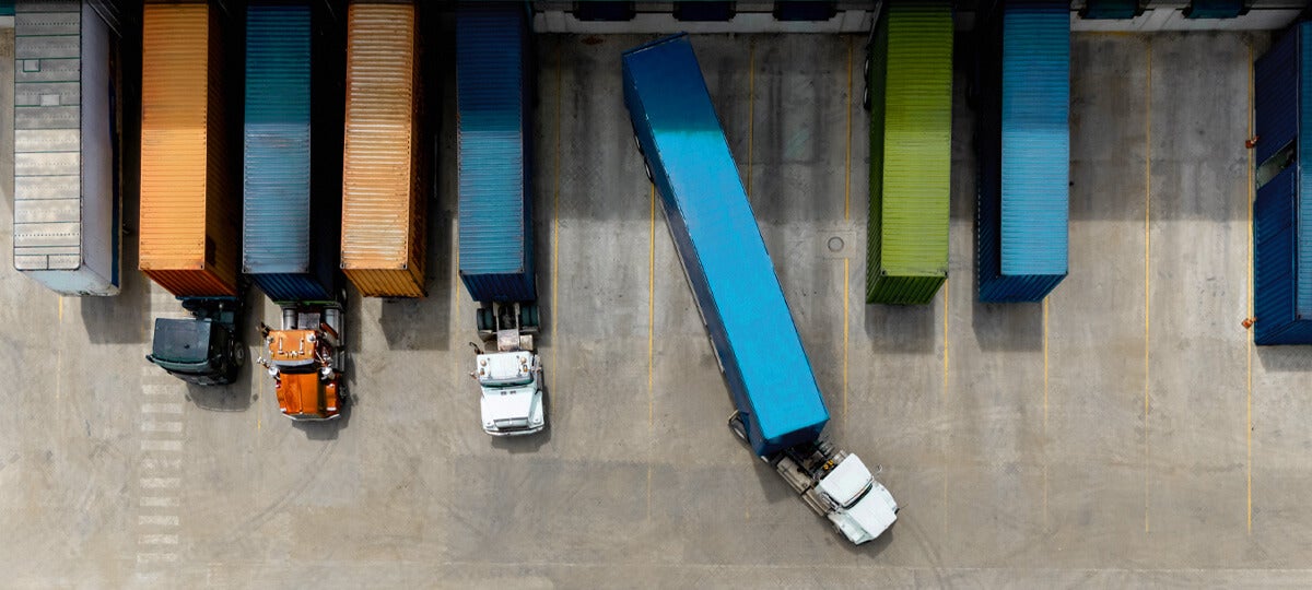 Aerial view of semi-trucks parked at a loading dock with colorful shipping containers highlights the precision of manufacturing logistics; one truck is backing up at an angle while others are parked straight.