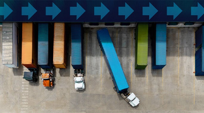 Aerial view of several trucks parked next to colorful shipping containers at a loading dock, streamlining manufacturing logistics. One truck maneuvers to park as blue arrows painted above the containers point right.