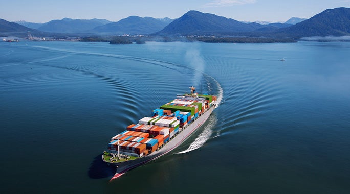 A large cargo ship loaded with colorful shipping containers sails through calm blue water, leaving a wake behind, symbolizing efficient parcel spend management amid forested mountains and a partially cloudy sky in the background.