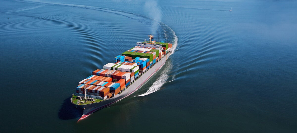 A large cargo ship carrying colorful shipping containers moves through calm blue water, optimizing logistics with parcel spend management, and leaves a broad wake behind it under a clear sky.