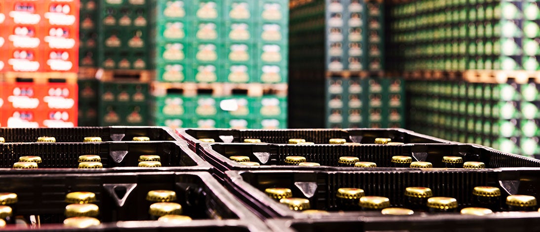 Close-up of crates filled with bottles capped with golden tops, set against blurred stacks of green and red crates in the background, highlighting a busy food and beverage supply chain warehouse or storage facility.